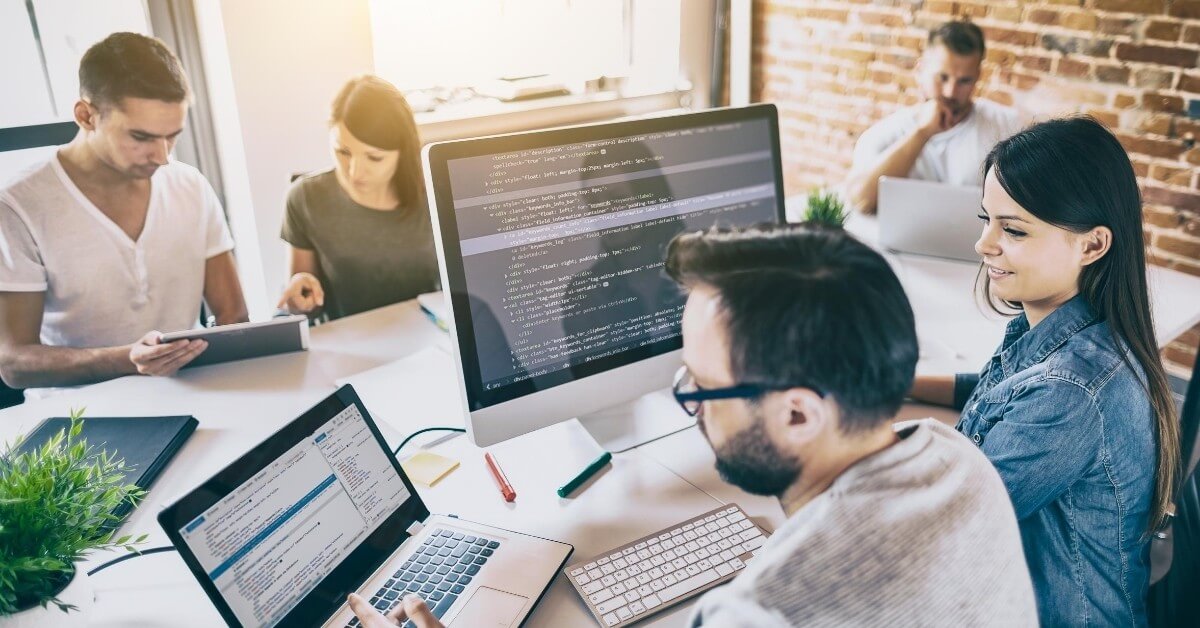 Woman sitting with a software developer at his desk with other coworkers in the background